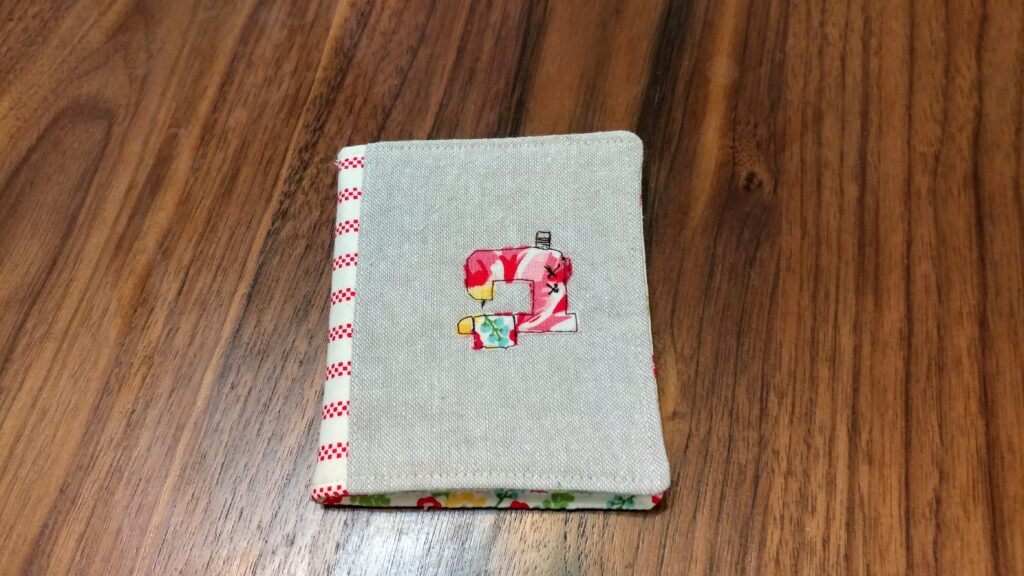 Front of a needlebook.  There is an appliqued design of a sewing machine on a linen background.